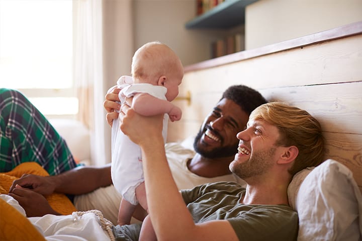 a mixed race gay couple holds up their adopted baby with joy
