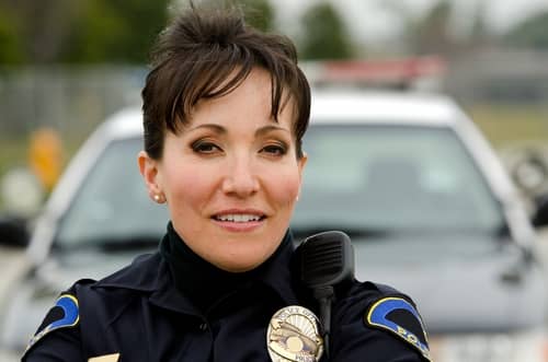 A female police officer in a dark colored uniform standing in front of a patrol car
